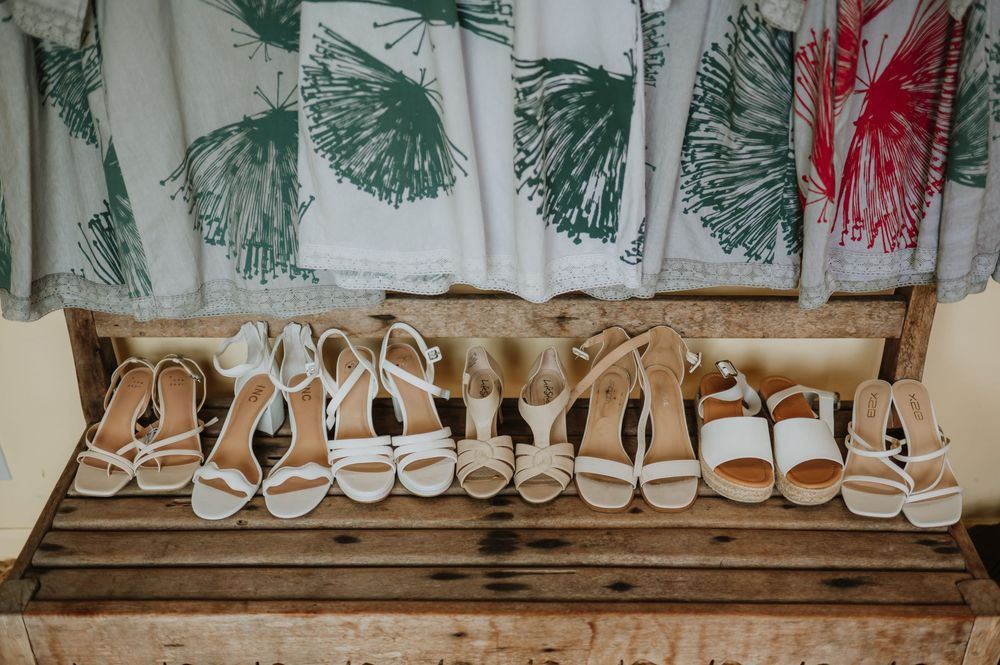 A row of elegant white bridal shoes and sandals arranged neatly on a wooden shelf with floral wallpaper background.