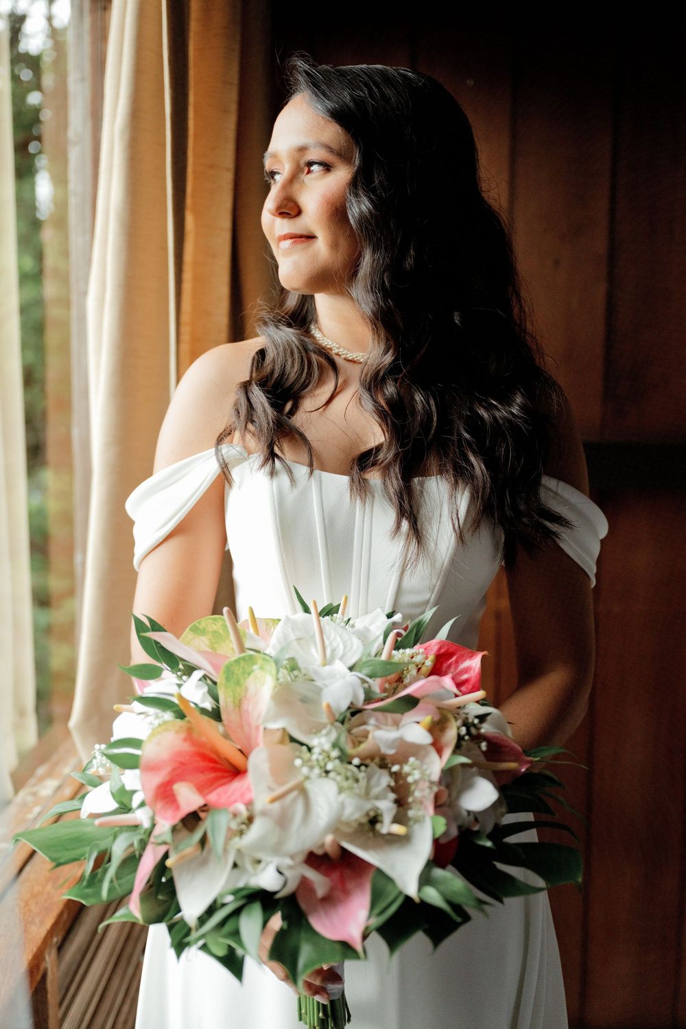 A person in a white dress holds a pink and white lily bouquet while gazing out a window in a rustic wooden setting.