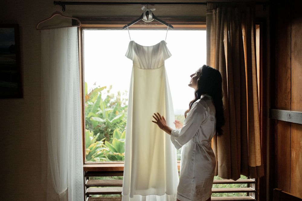 A white wedding dress hangs by a window with soft natural light streaming through sheer curtains.