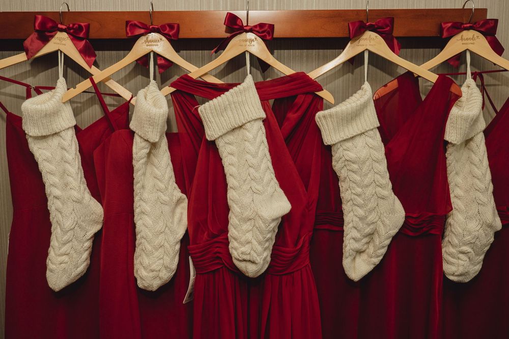 Red velvet dresses and white knit Christmas stockings hanging on wooden hangers against a brown wall.