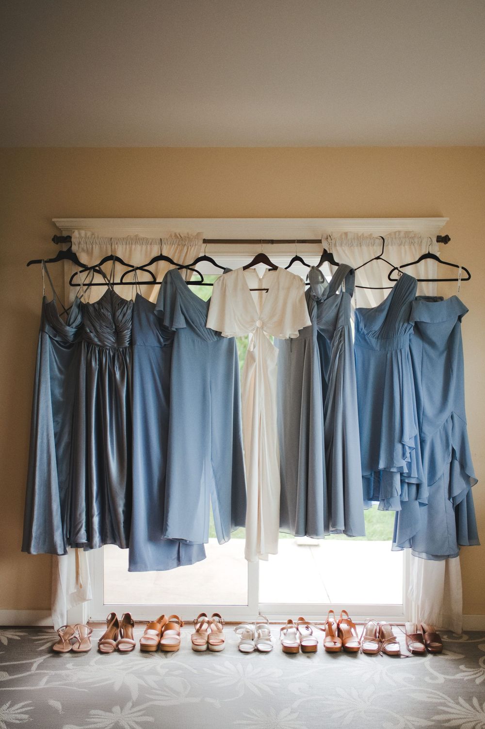 Row of blue bridesmaid dresses hanging on curtain rod with matching nude shoes lined up beneath them.