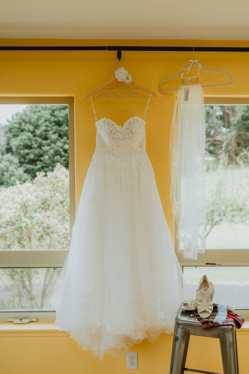 A beautiful white wedding dress with a sweetheart neckline and lace details hangs by a window in a yellow room.