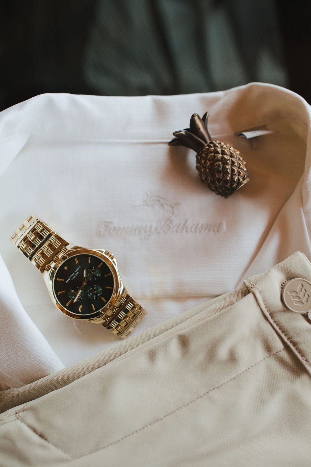 Gold wristwatch with black face displayed on white fabric with small decorative pineapple accent.