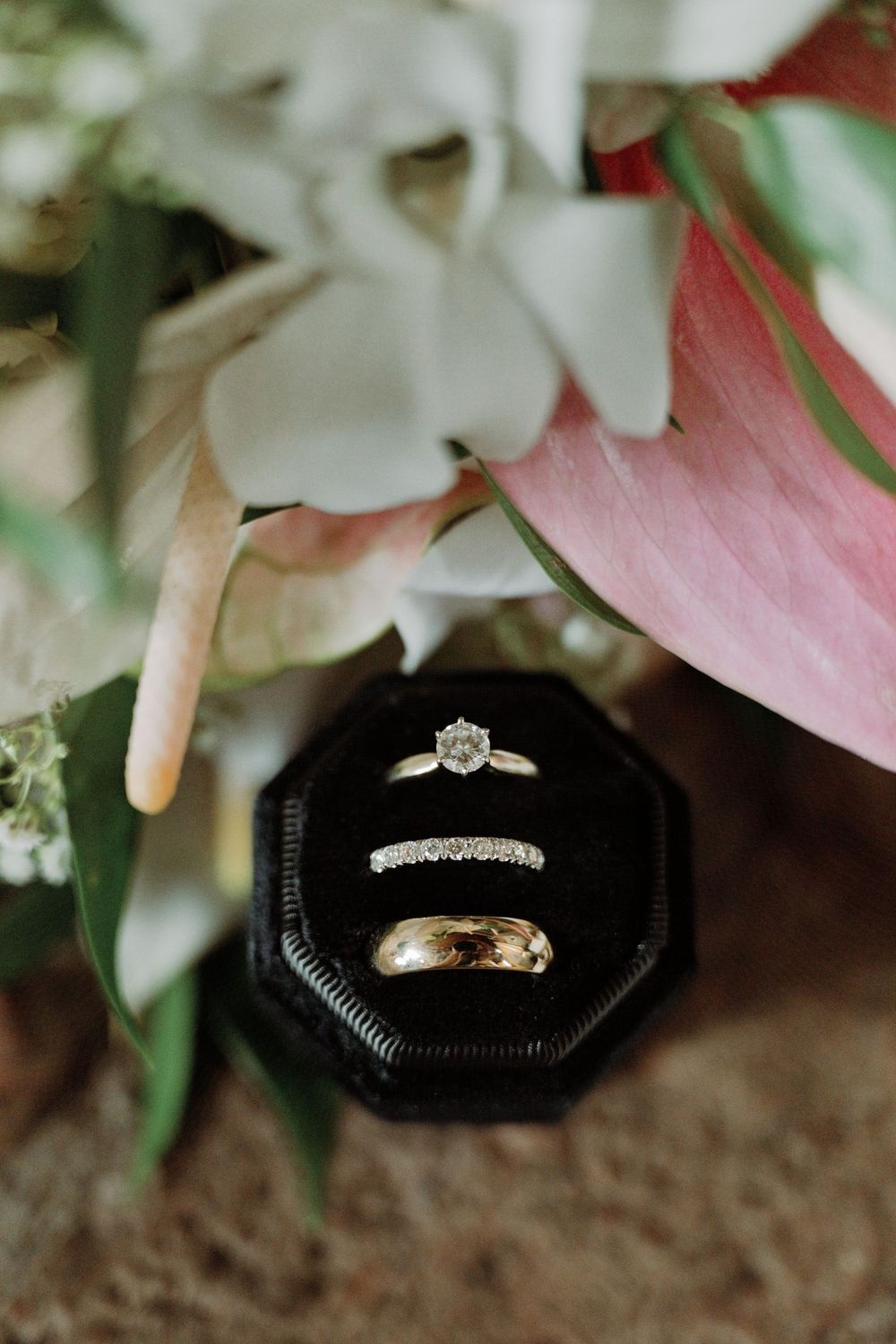 Wedding rings and engagement ring displayed in a black octagonal box surrounded by soft pink and white flowers.