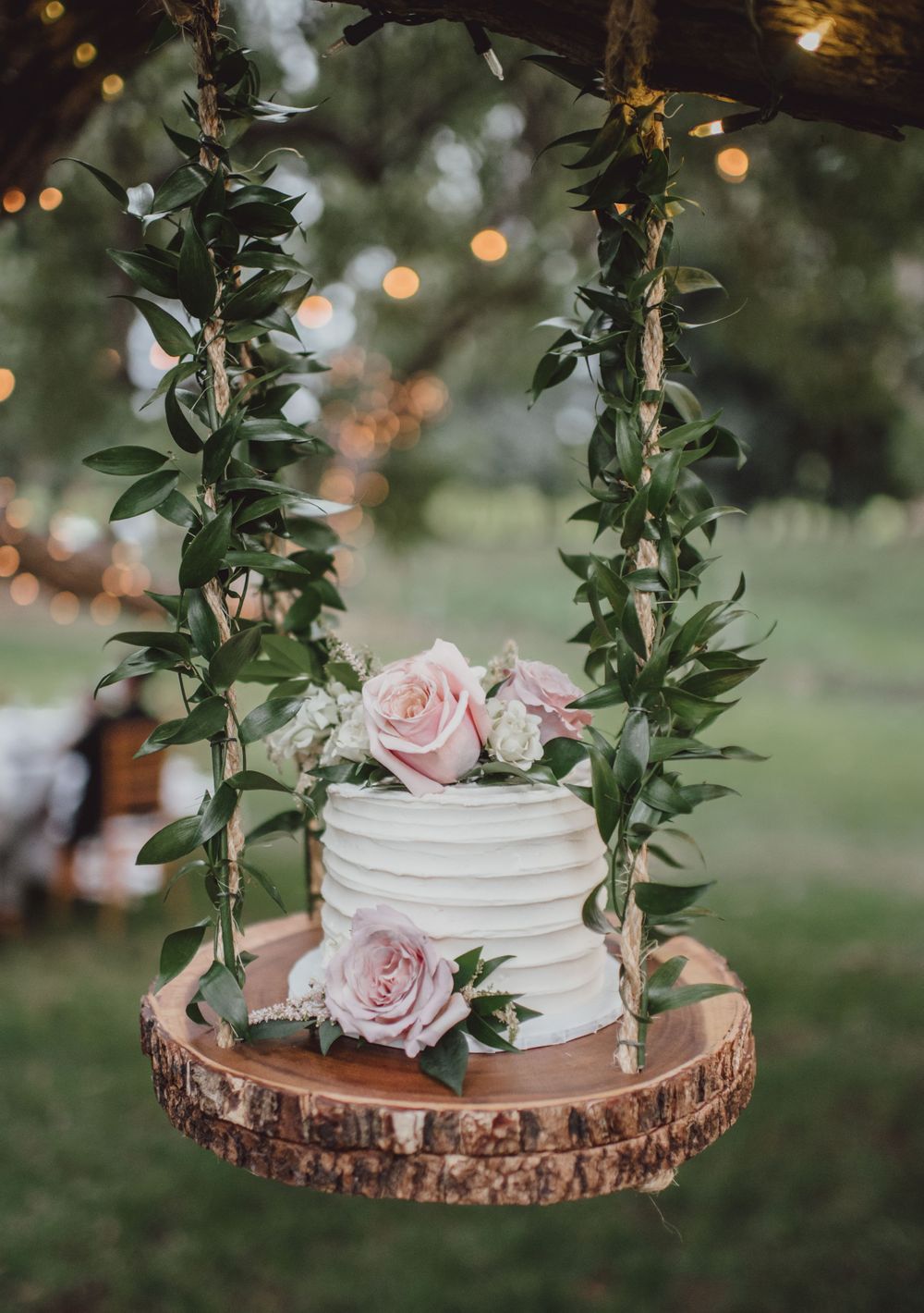 White wedding cake with pink roses sits on rustic wood slice with greenery garland decoration.