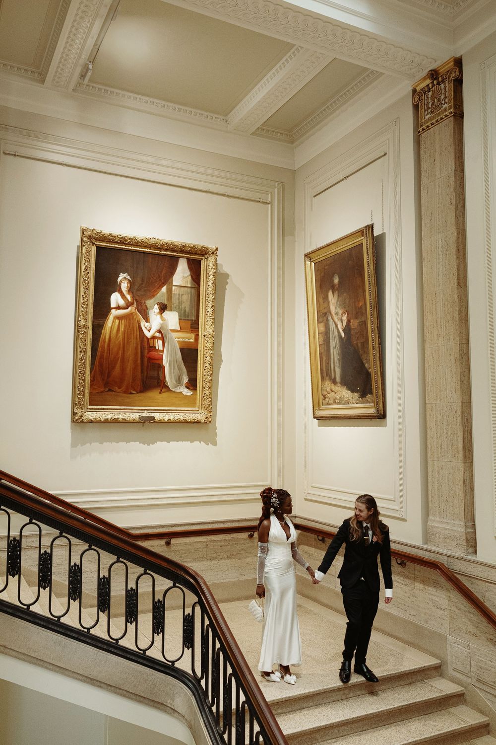 A couple ascending an elegant white staircase with ornate railings and classical paintings on the walls.