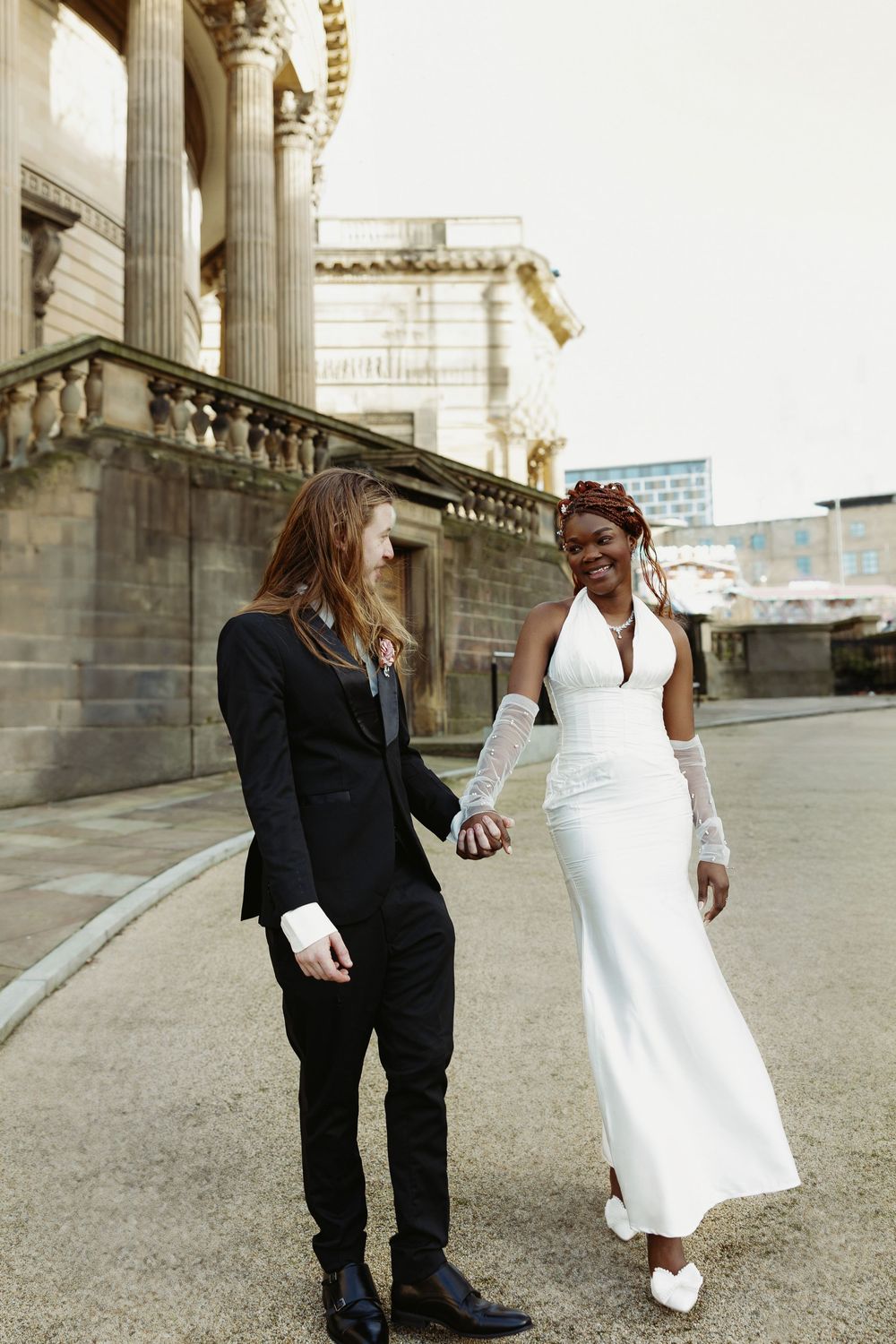 A couple in formal attire, with a white dress and black suit, share playful moments outside a classical building.