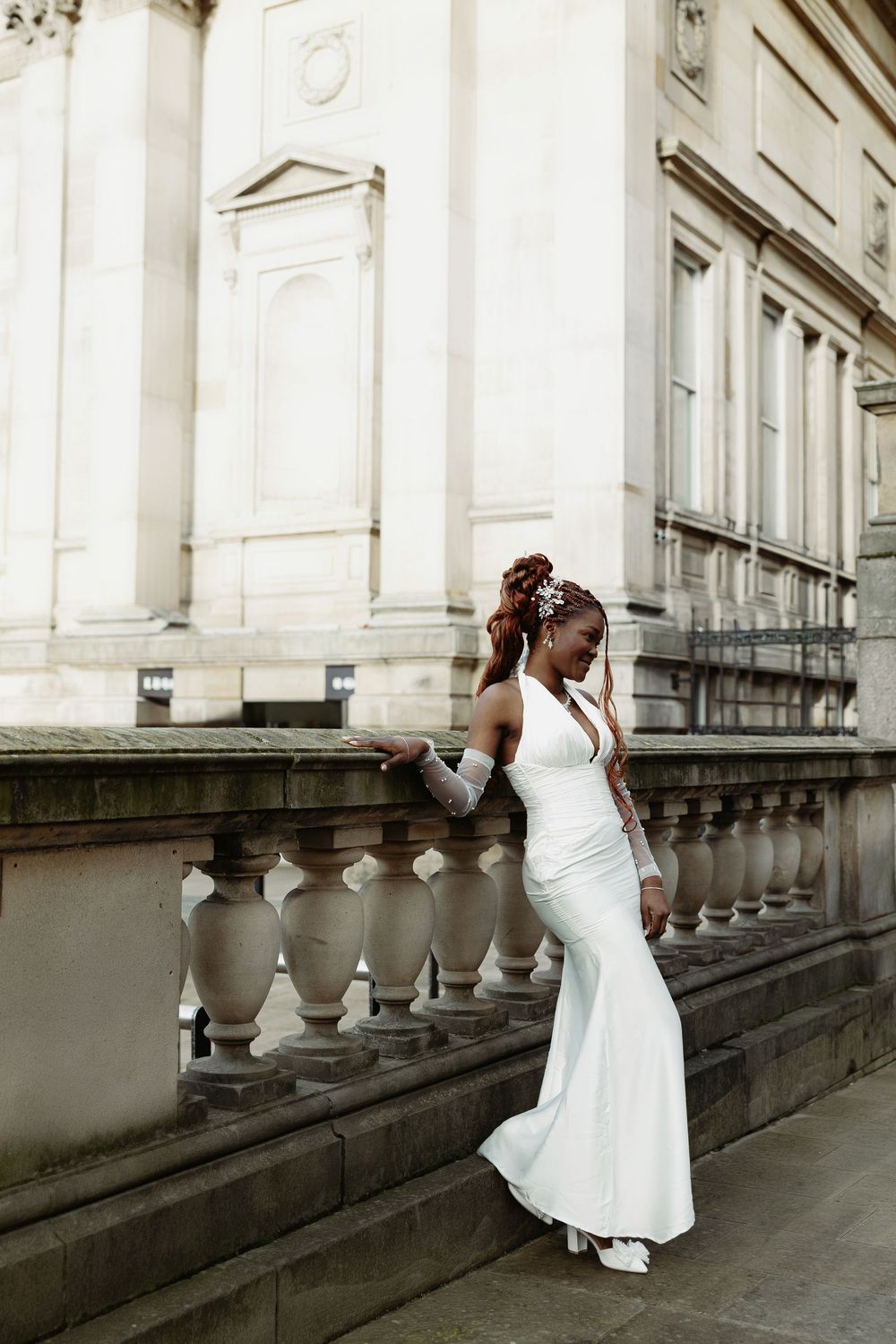 A person in a white gown poses on grand stone steps of a historic building with ornate balustrades.