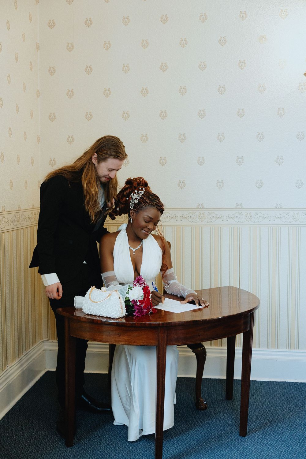 Two people share a moment at an antique wooden table during a wedding ceremony signing in a room with vintage wallpaper.