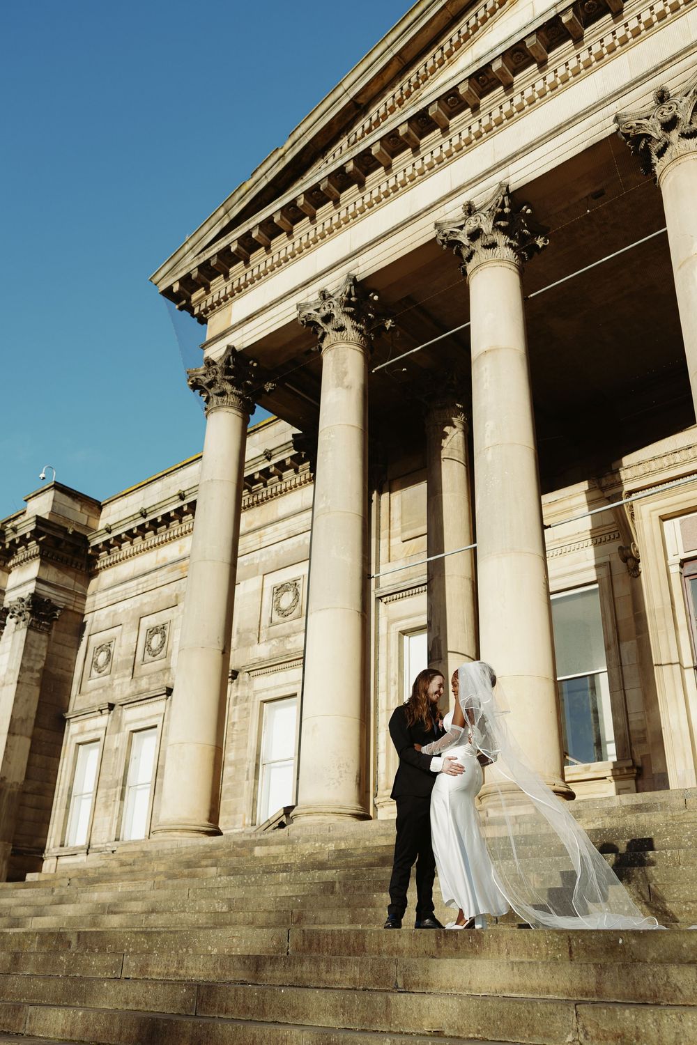 A couple shares a romantic moment on the steps of a neoclassical building with towering columns.