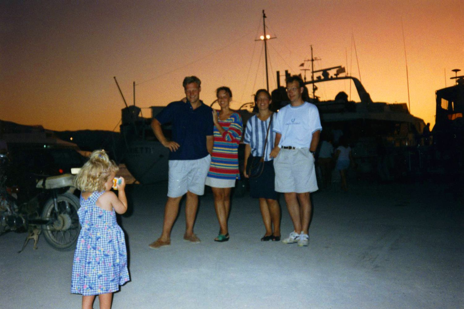 Lite barn som tar bilde av 4 voksne med kameraet sitt. Sola er på vei ned og bak den ser vi konturer av seilbåter ved en brygge.