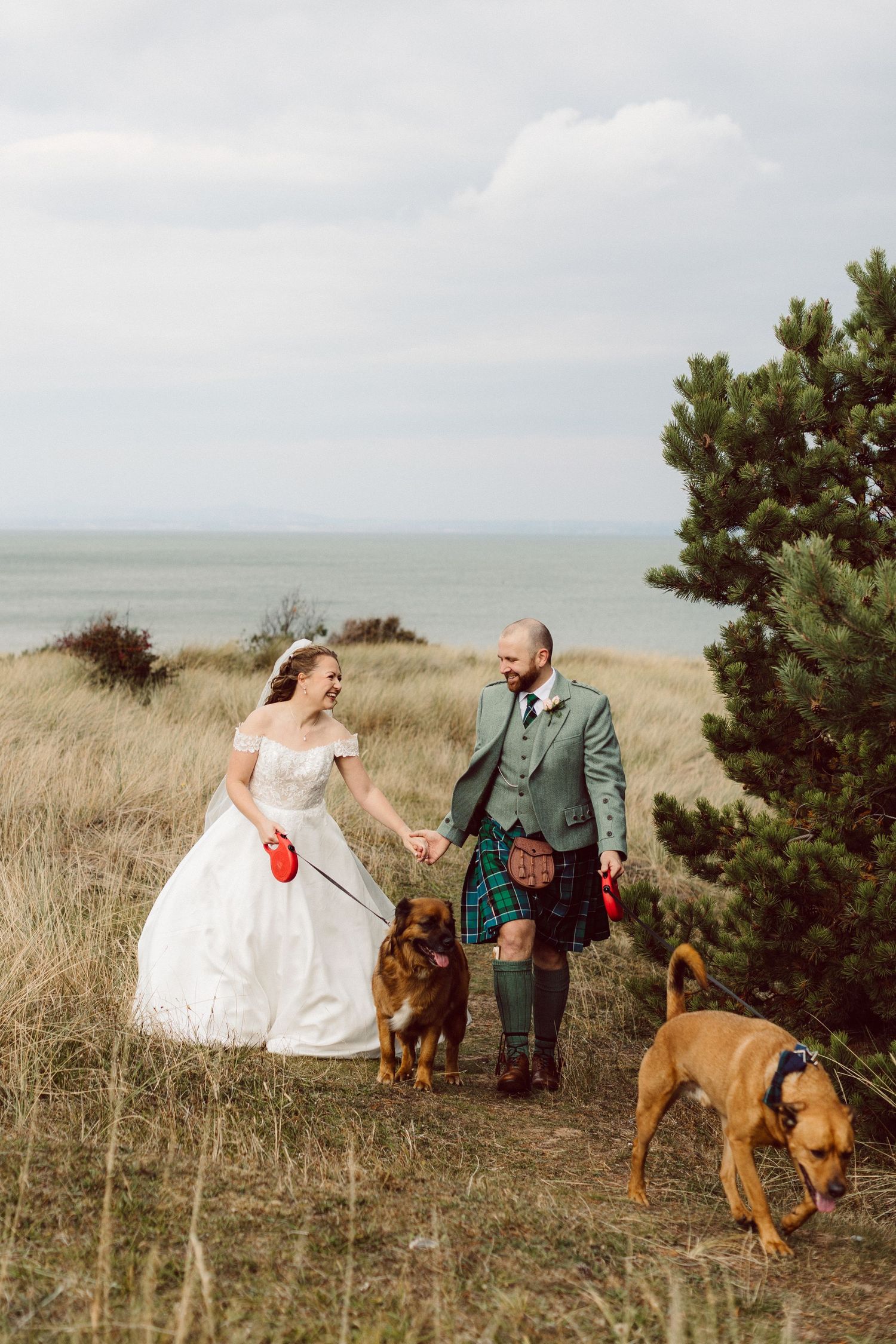 A couple in wedding attire walks along a coastal path with their dogs against an overcast sky and ocean backdrop.