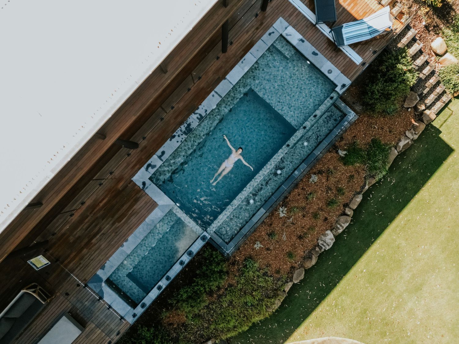 Aerial view of a person floating in a rectangular luxurious backyard pool surrounded by wooden decking and landscaped gardens.