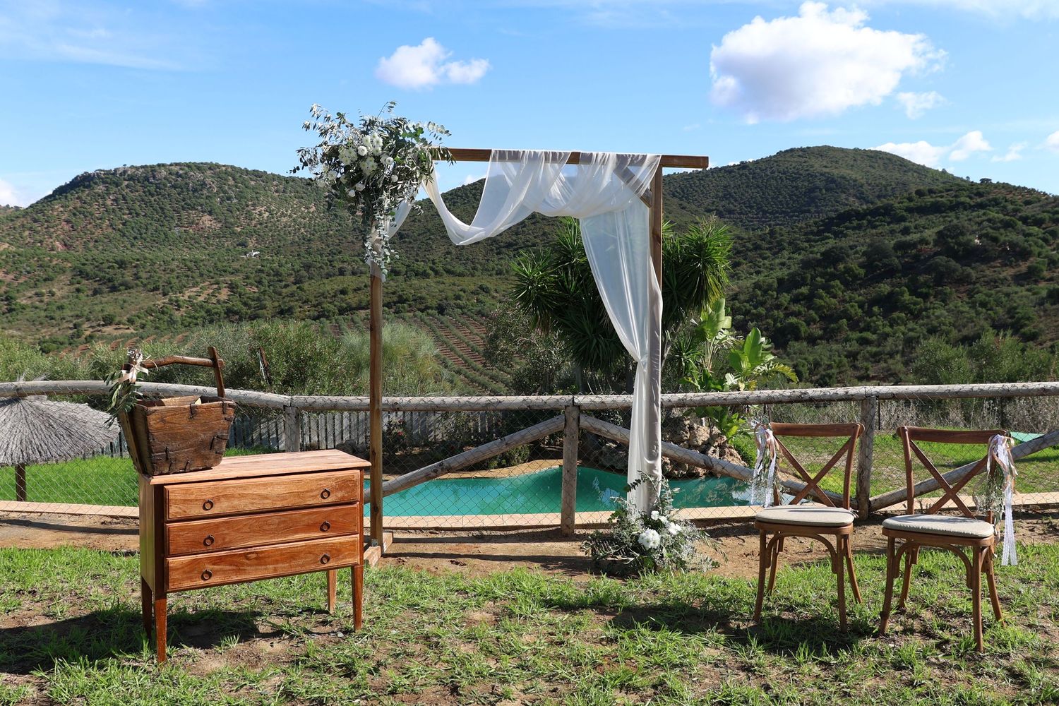 A scenic outdoor patio with wooden furniture and white curtains overlooking green mountains and a swimming pool.