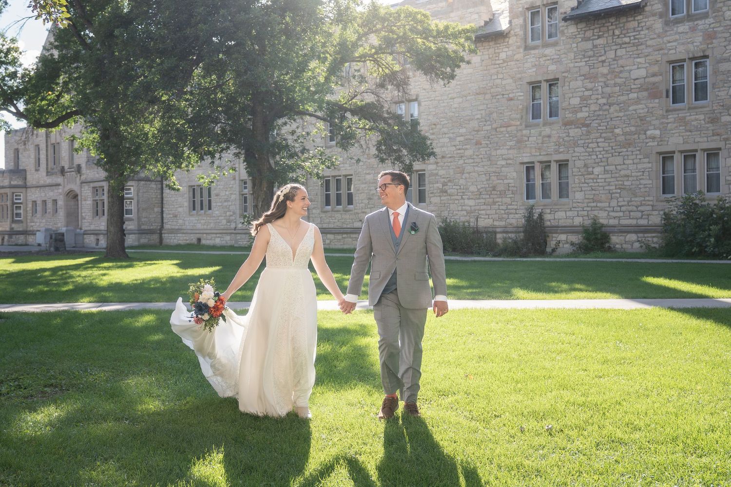 A couple walks hand in hand across a sunny lawn outside a stone building during a wedding celebration.