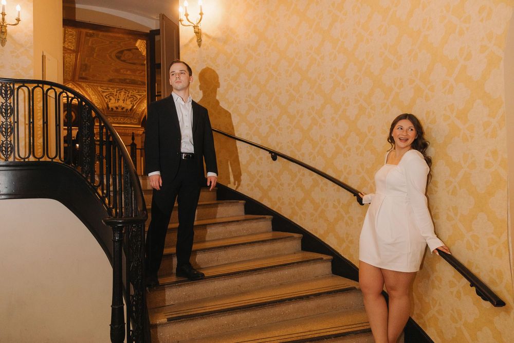 A wedding couple descends an elegant staircase with yellow wallpaper and ornate lighting fixtures in a grand venue.