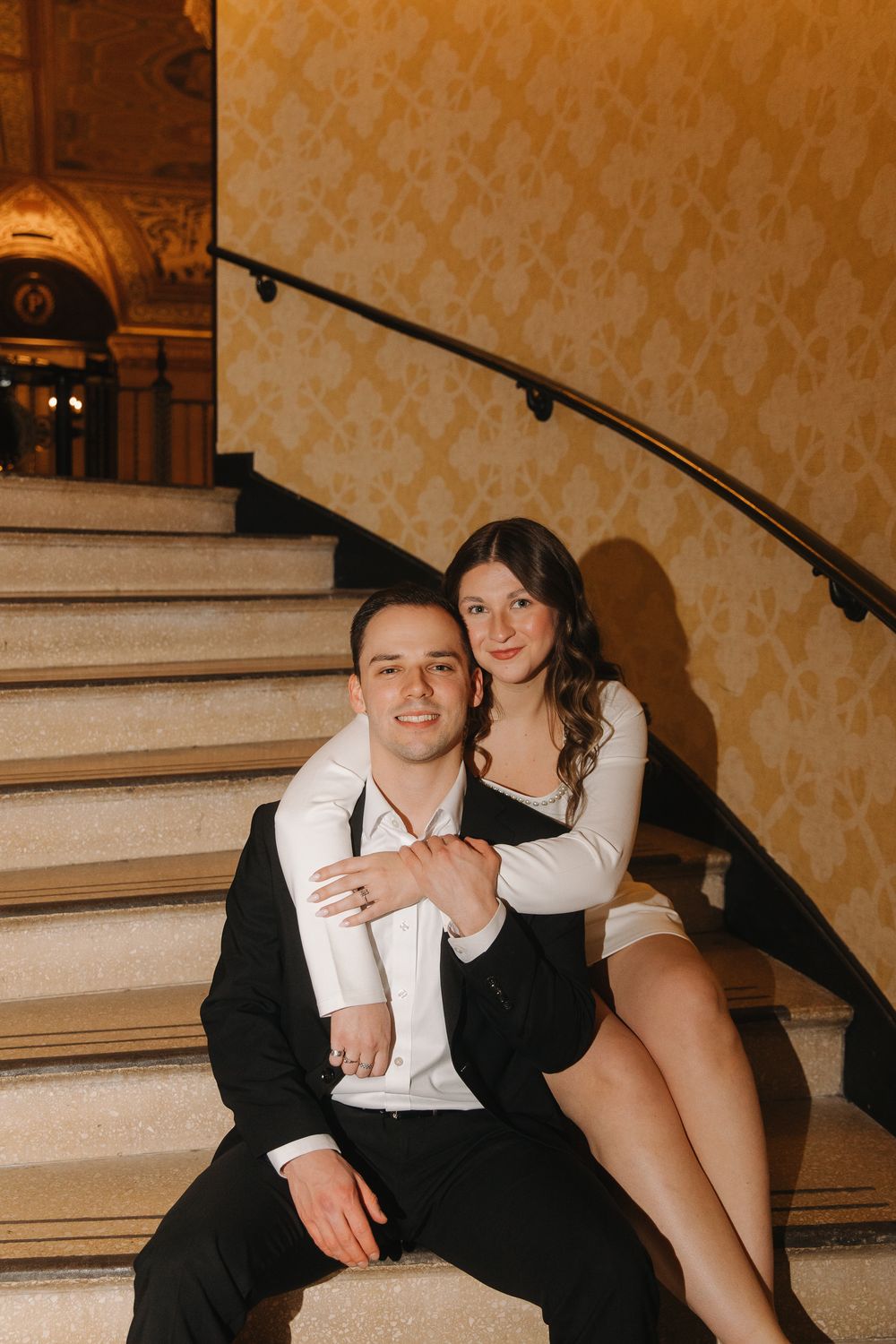 A couple in formal attire poses together on an elegant staircase with patterned wallpaper in the background.