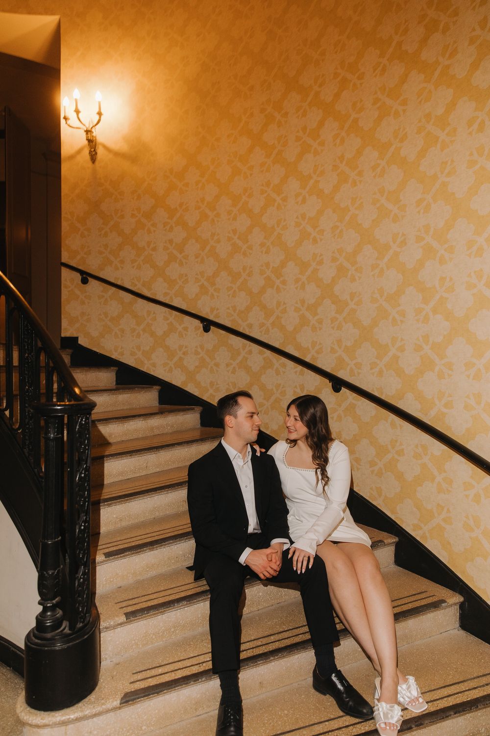 A romantic couple sits together on elegant stairs with yellow patterned wallpaper in a vintage-style interior setting.