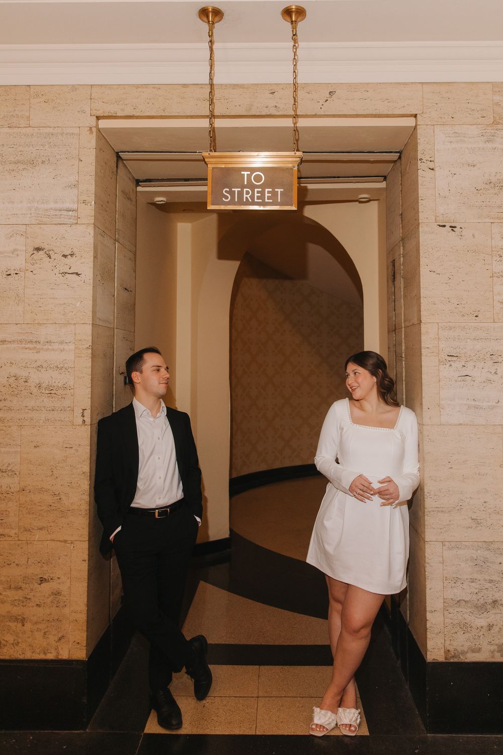 A couple in black and white outfits poses playfully in a vintage hallway with cream-colored walls.