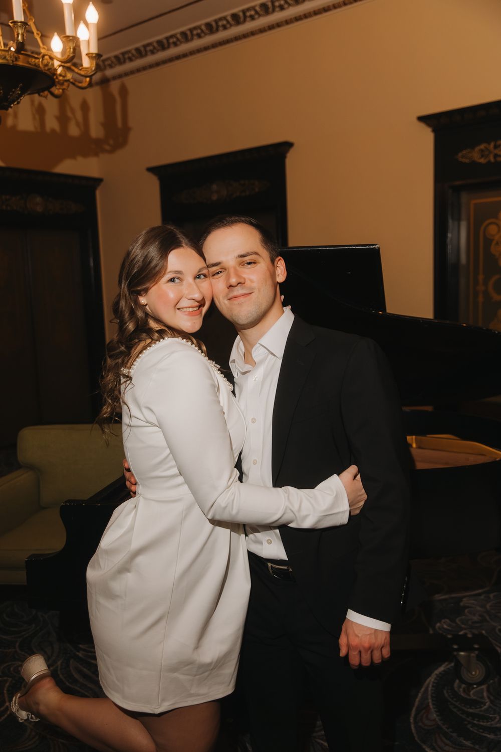 A couple in formal attire embrace in a warmly lit room with elegant decor and a chandelier overhead.
