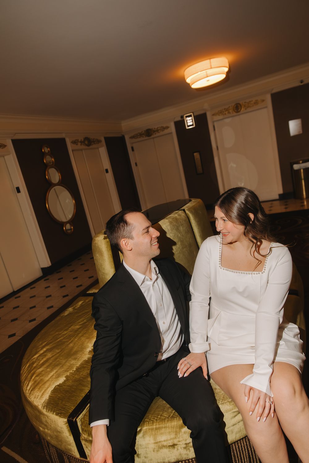 A couple in formal attire sits together on a gold velvet couch in an intimate moment.