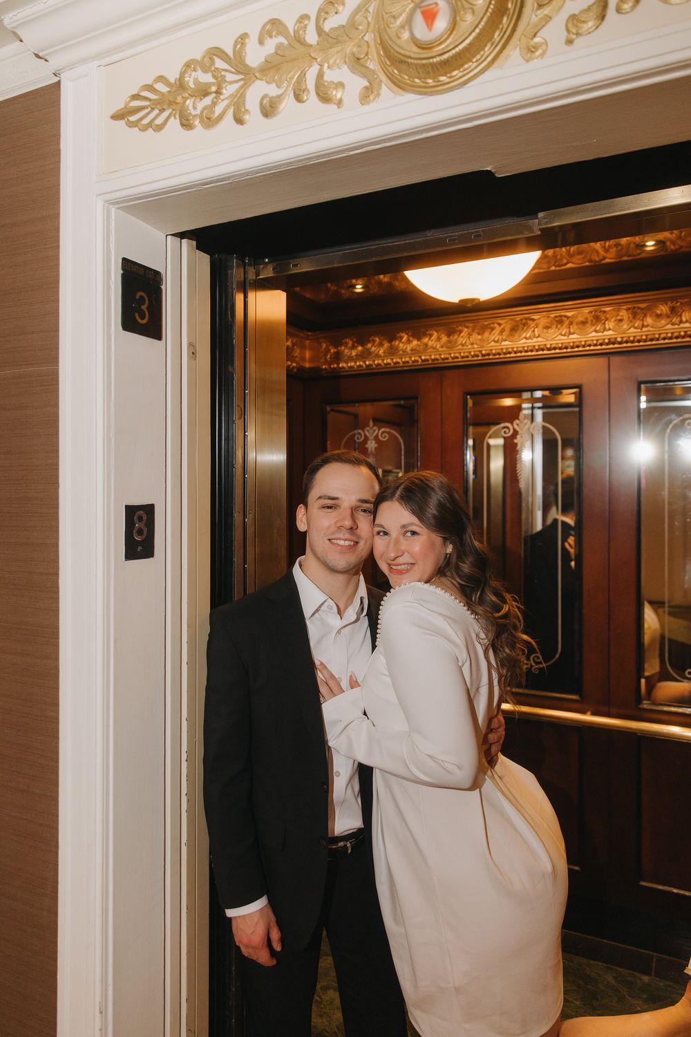 A couple shares a joyful moment in an elegant hallway with ornate gold trim and warm lighting.