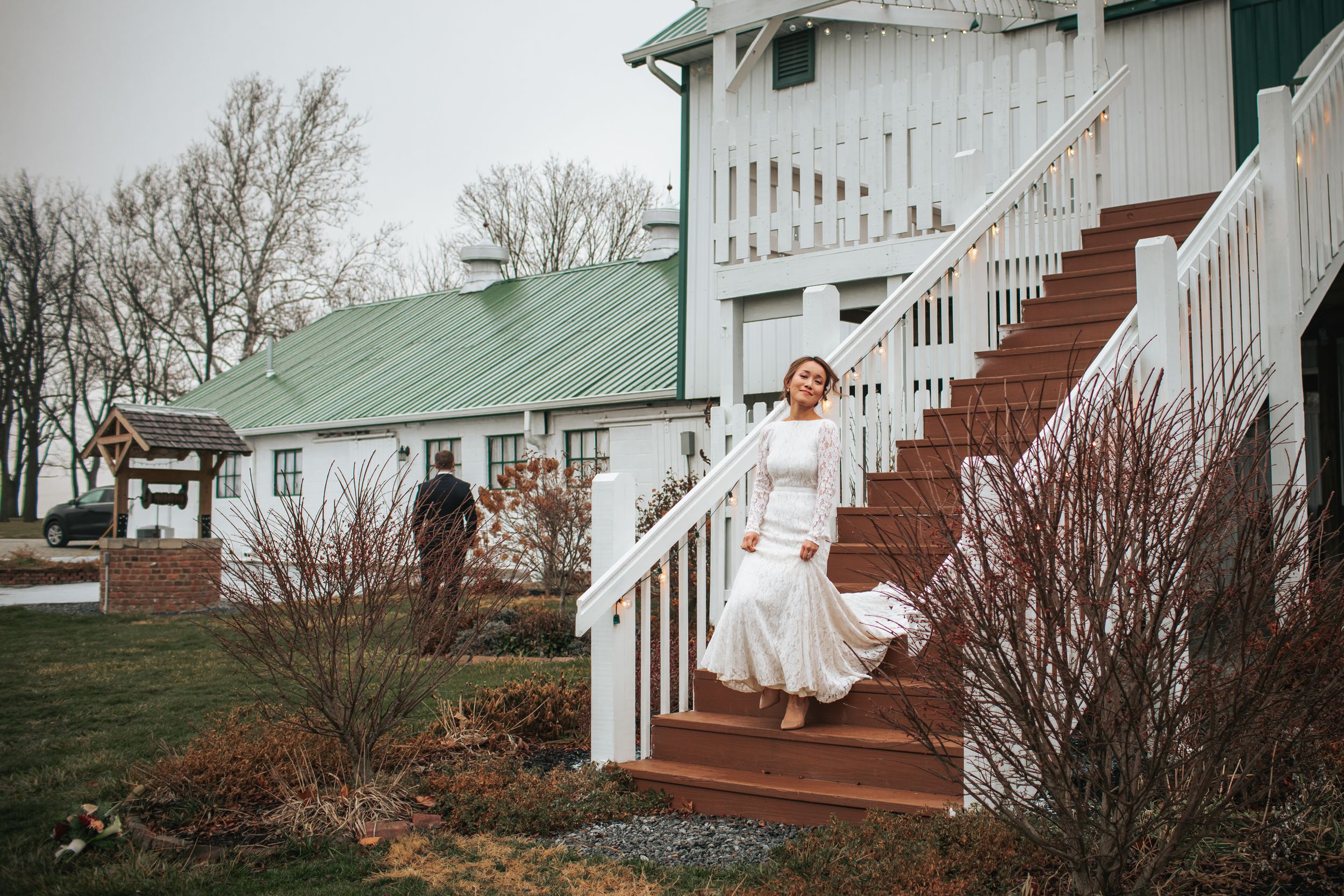 A white wedding dress flows down wooden stairs at a rustic venue with a green metal roof and white railings.