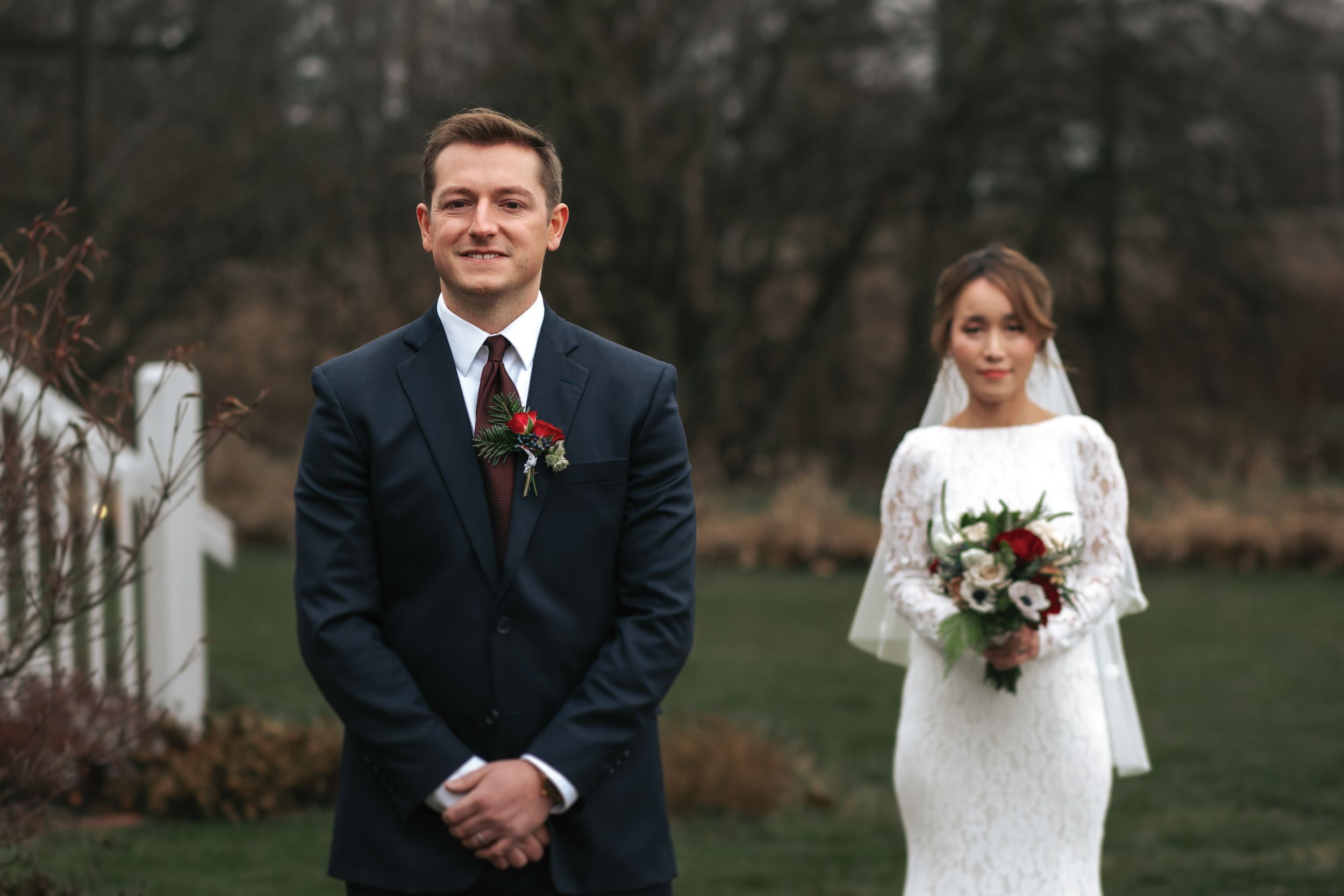 A couple in wedding attire stands together in a garden setting during an autumn day.
