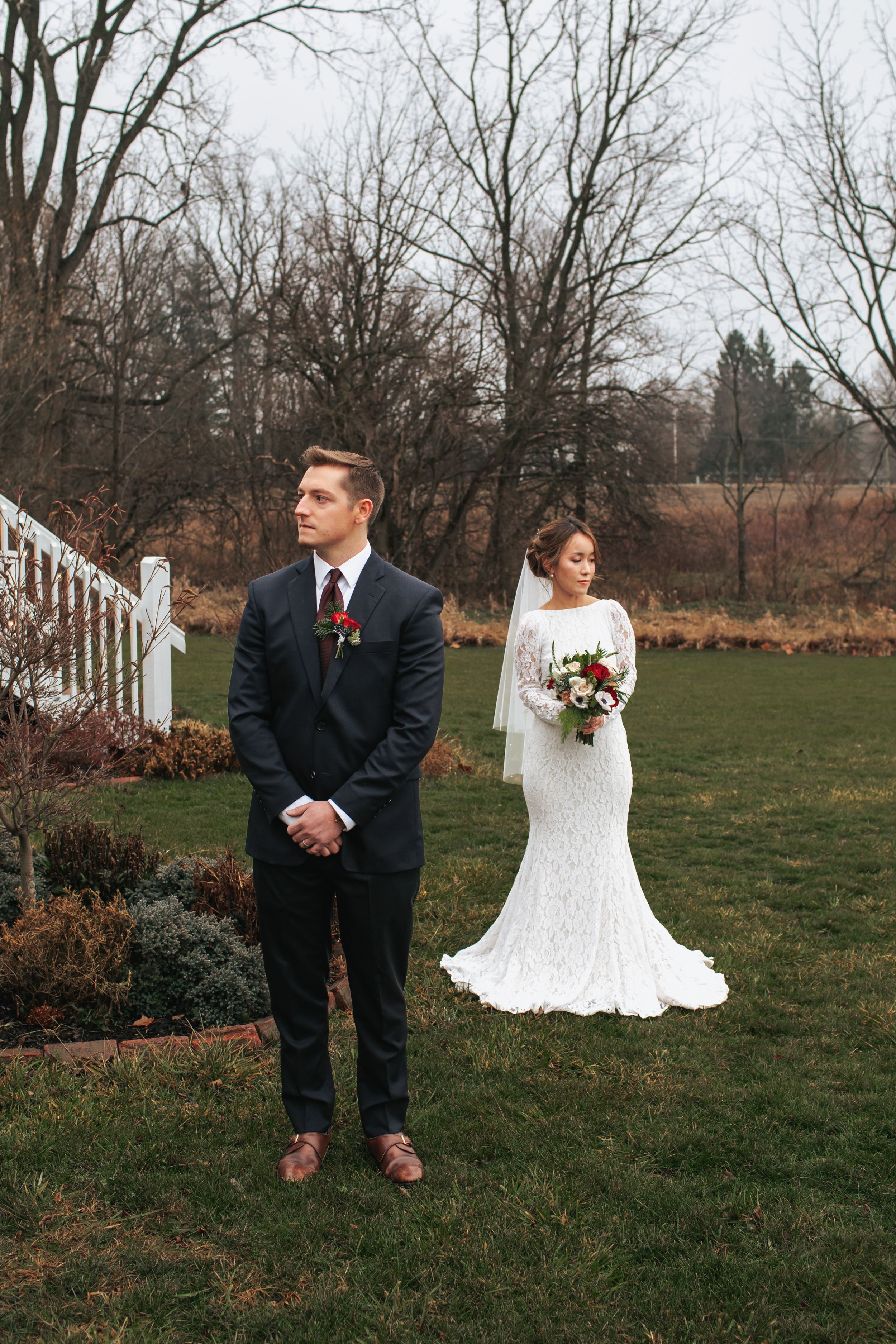 A bride and groom stand apart during their first look moment in a garden setting on an overcast winter day.