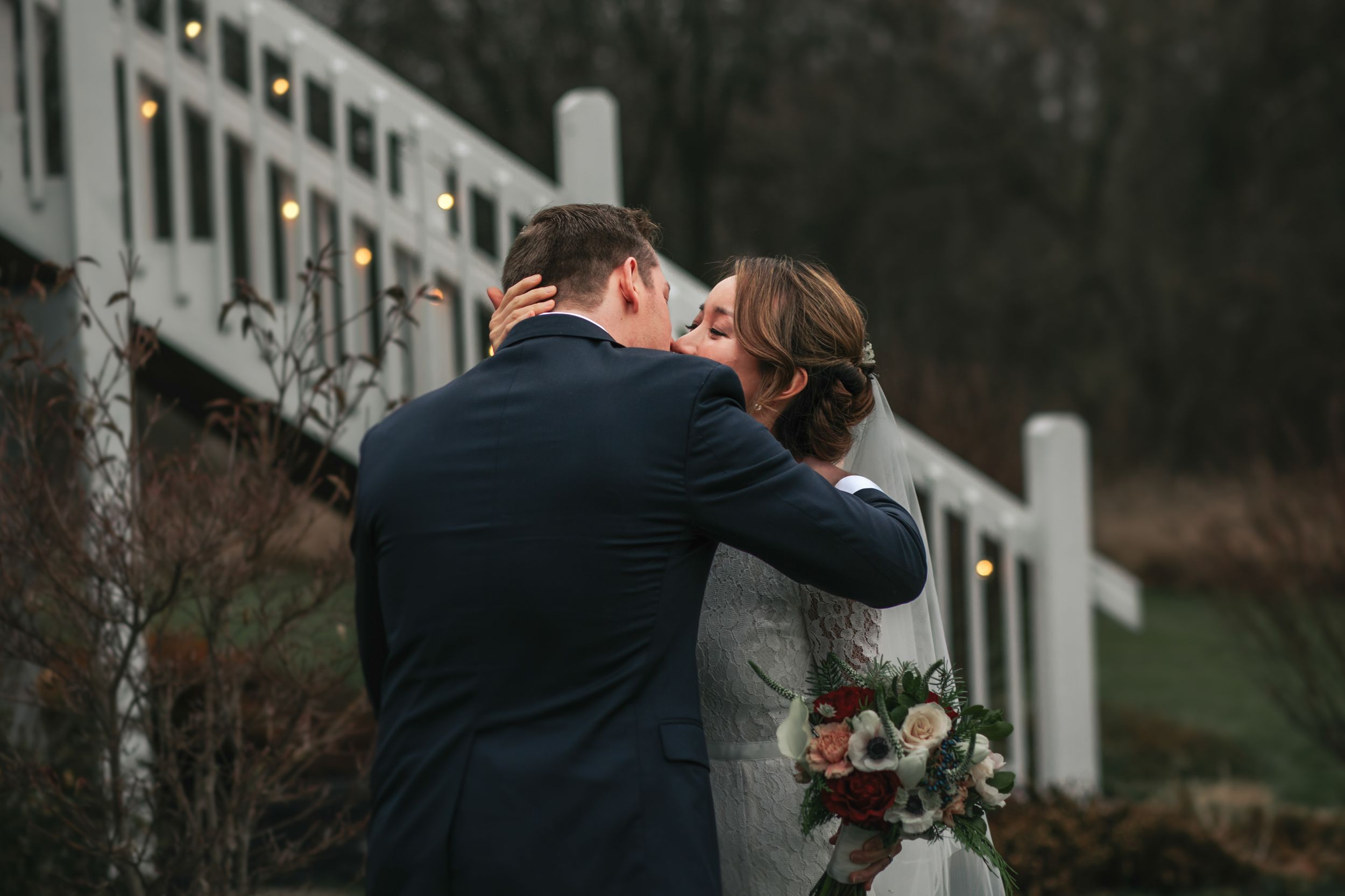 A romantic couple shares a tender kiss on a bridge surrounded by white railings and autumn trees in the background.