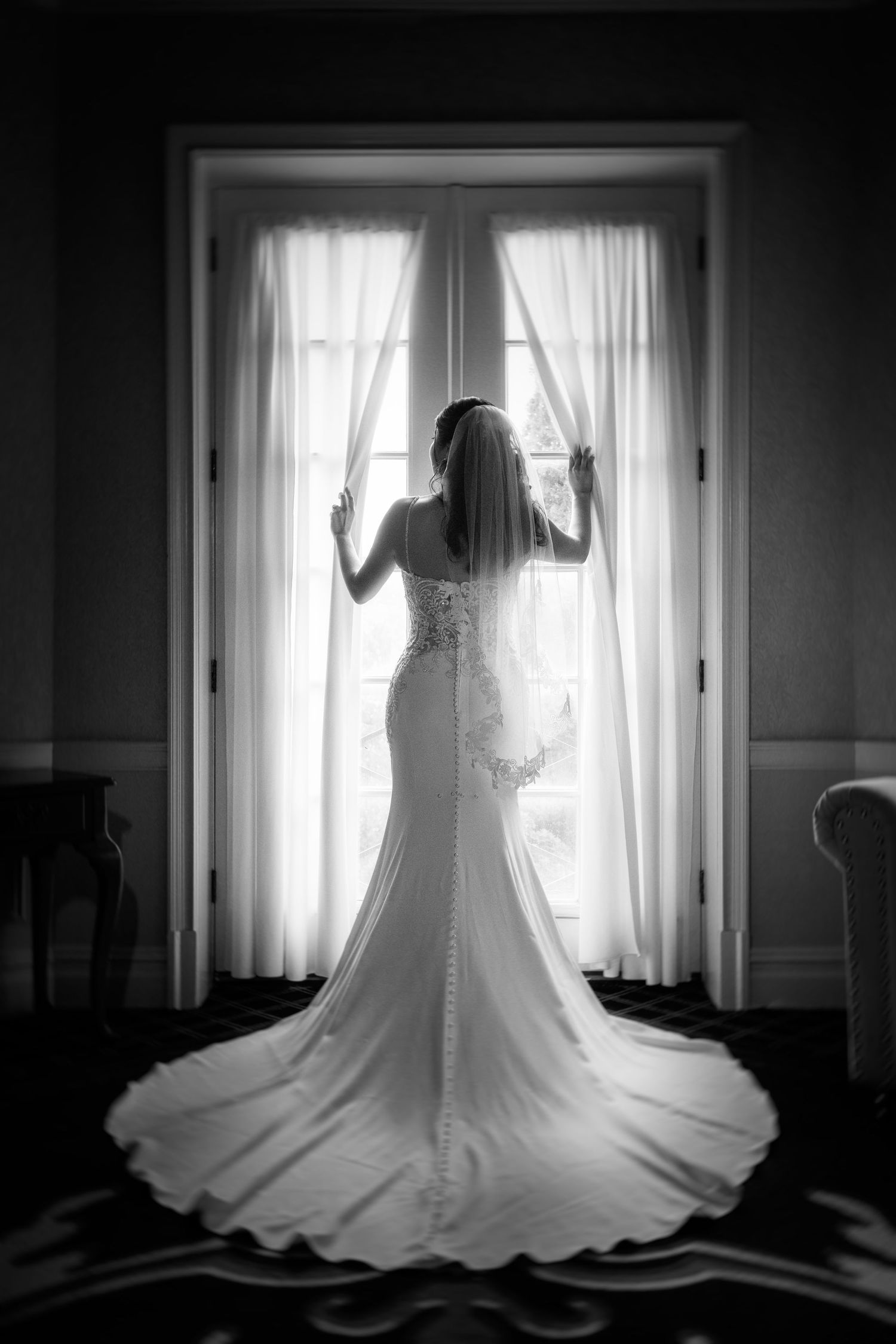 A dramatic black and white silhouette of a bride in a fitted lace gown by a window.