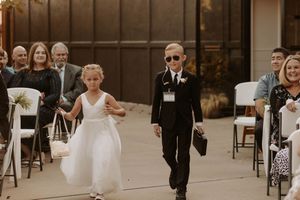 A wedding party walks down a brick-paved street in formal attire with bridesmaids in black dresses and flower girls in white.