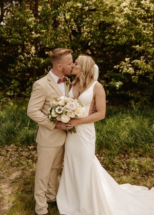 A couple in wedding attire share romantic kisses in a lush outdoor garden setting with natural sunlight filtering through trees.