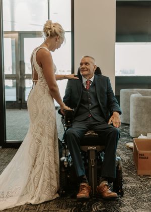 A person in a white lace wedding dress stands next to someone seated in a wheelchair wearing a dark suit.