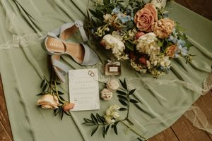 Delicate wedding details arranged on sage green fabric including flowers, invitations, rings, and perfume bottles.