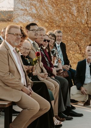 A row of wedding guests seated on wooden benches during an outdoor ceremony in warm autumn lighting.