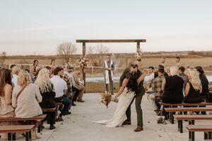 Wedding ceremony guests gather at rustic wooden benches in an outdoor venue at sunset.