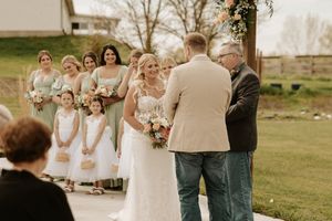 A rustic outdoor wedding ceremony with bridesmaids in blue dresses and floral bouquets on a grassy field.
