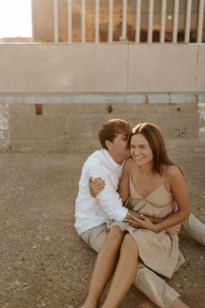 A romantic couple shares intimate moments while sitting on concrete ground during a sunset photoshoot.