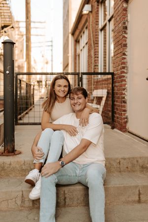A couple sits casually on concrete steps against a brick wall in matching white tops and light denim.