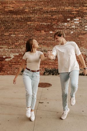 Two people walking together in coordinated outfits of white tops and light jeans along a brick wall.
