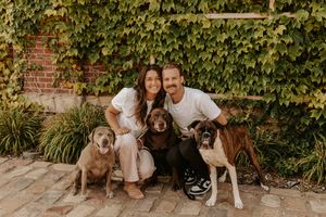 A group poses with their three dogs in front of an ivy-covered brick wall in an outdoor garden setting.