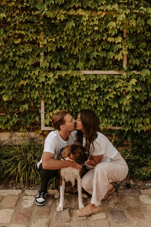 A couple shares a tender moment with their dog against a wall covered in lush green ivy.