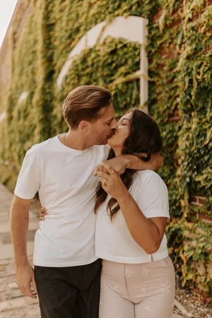 A romantic couple in matching white t-shirts share tender moments against an ivy-covered wall in warm sunlight.