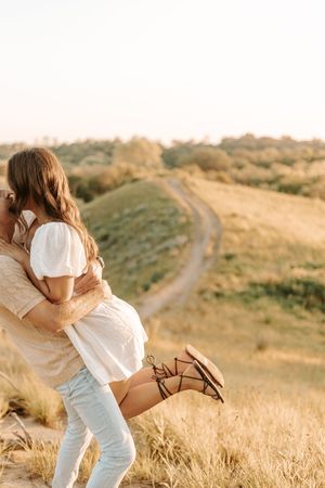 A couple in white clothing embraces tenderly in a golden field at sunset with winding paths in the background.