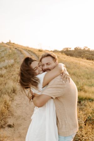 Couple embraces lovingly in golden field during sunset while wearing neutral toned outfits.