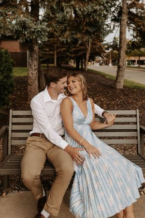 A romantic couple sits together on a wooden park bench surrounded by trees in an autumn setting.