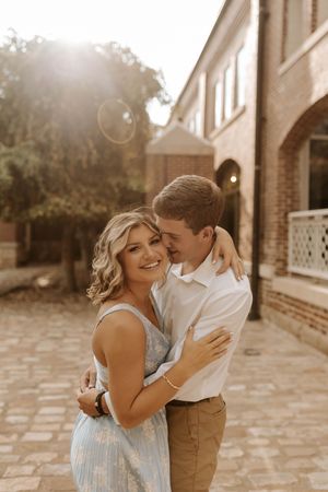 A romantic couple embraces on a brick patio in warm sunlight wearing formal attire.