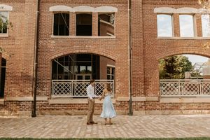 A couple shares a romantic moment in front of a historic brick building with arched windows and decorative railings.