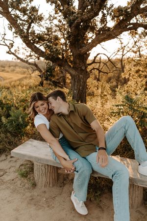 A couple shares a romantic moment while sitting on a wooden bench under a large tree at sunset in a golden field.